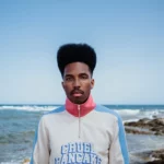 Jeune homme noir portant un pull blanc avec des bandes bleues sur les manches, un col montant rouge et zippé, avec le logo Cruel Pancake bleu au centre