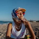 Jeune femme portant un marcel blanc avec l'inscription Cruel Pancake en orange et des illustration de Lipton et Cruel Pancake sur le coeur. Elle porte une casquette recouverte d'un foulard coloré, noué sous le menton. La plage et la mer en fond. Une cannette de Lipton Ice Tee posée devant elle.